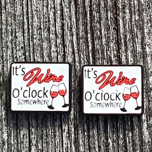 May include: Two rectangular charms with the text "It's Wine O'clock Somewhere" in red and black. The charms feature two wine glasses with red wine. The background is a weathered wood.