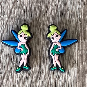 May include: Two Tinkerbell shoe charms, each featuring a cartoon image of Tinkerbell with green wings, a green dress, and yellow hair. The charms are made of rubber and have a blue and green color scheme.