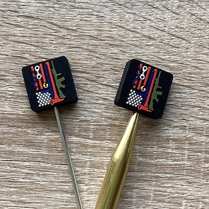 May include: Two black square-shaped knitting needle tips with a colourful design of an American flag with guns, handcuffs, and a wrench. The tips are attached to two knitting needles, one gold and one silver.