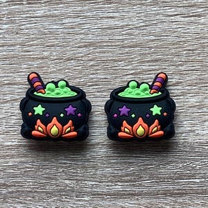 May include: Two black cauldron charms featuring a bright green bubbling liquid, a striped stirring stick, and orange flames at the base. The exterior of each cauldron is decorated with small purple and green stars against the black surface.