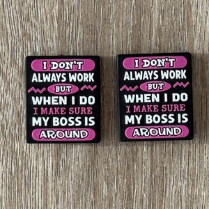 May include: Two rectangular black charms featuring pink and white text that reads: I DON'T ALWAYS WORK BUT WHEN I DO I MAKE SURE MY BOSS IS AROUND. The design includes pink zig-zag accents and pink oval borders around specific words on a flat surface.