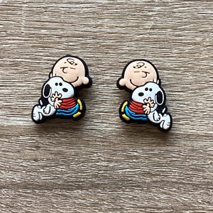 May include: Two identical shoe charms featuring a colorful graphic of the cartoon character Charlie Brown hugging his dog Snoopy. The figures are rendered in a flat, rubberized style with black outlines, wearing a red shirt and blue pants.