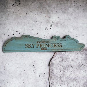 May include: A wooden cutout of a cruise ship in the shape of the Sky Princess cruise ship. The cutout is painted teal blue and has the text "November 2024 SKY PRINCESS Alaska" on it.