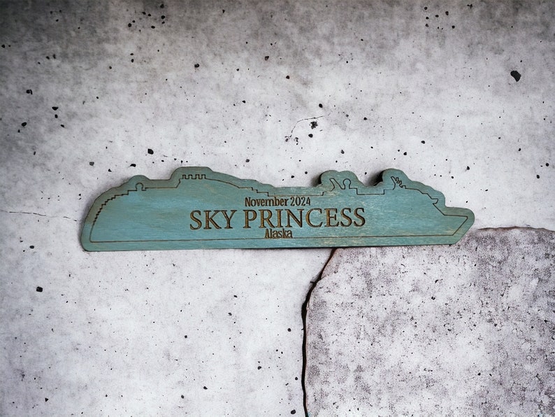 May include: A wooden cutout of a cruise ship in the shape of the Sky Princess cruise ship. The cutout is painted teal blue and has the text "November 2024 SKY PRINCESS Alaska" on it.