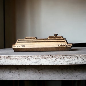 May include: A wooden ship ornament with the words "March 2022" and "Celebration" etched into the wood. The ship features a detailed design with multiple windows, and a light wood colour contrasting with the darker edges.