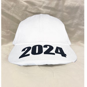 May include: A white baseball cap with the year "2024" printed in bold black letters across the front. The cap is made of a smooth, solid white material.