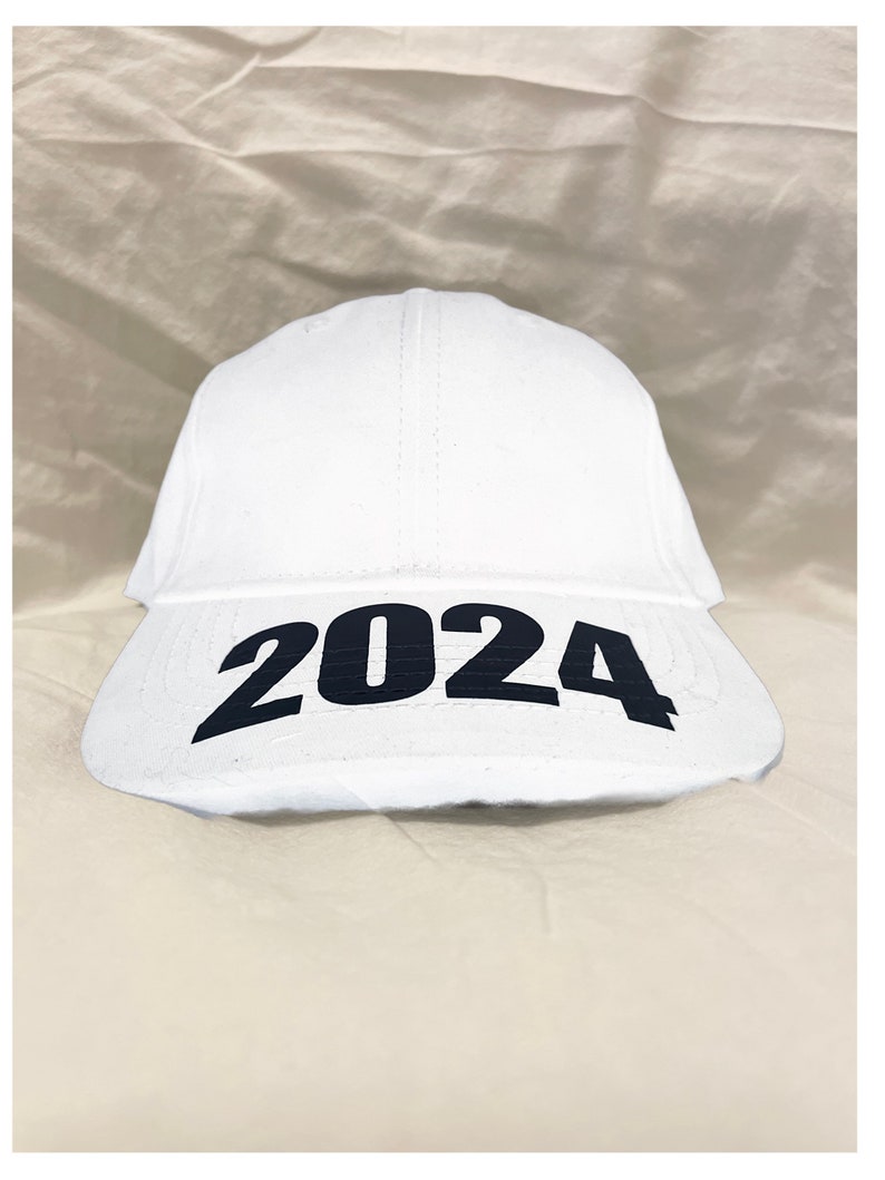 May include: A white baseball cap with the year "2024" printed in bold black letters across the front. The cap is made of a smooth, solid white material.