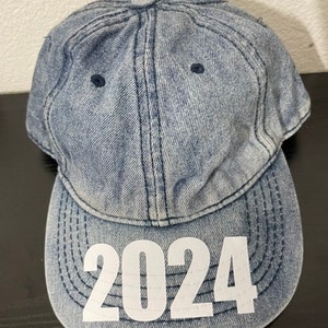 May include: A denim baseball cap with the year "2024" printed in white on the front. The cap is a light blue wash with a curved brim and a button on top. The stitching is visible.