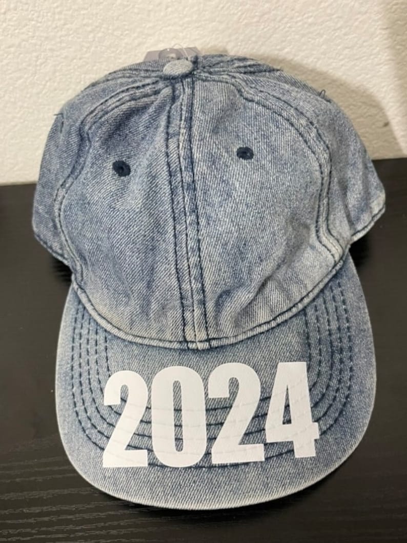 May include: A denim baseball cap with the year "2024" printed in white on the front. The cap is a light blue wash with a curved brim and a button on top. The stitching is visible.