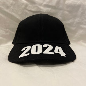 May include: Black baseball cap with the white text "2024" across the front. The cap has a curved brim and a structured crown. The hat is made of a dark fabric.