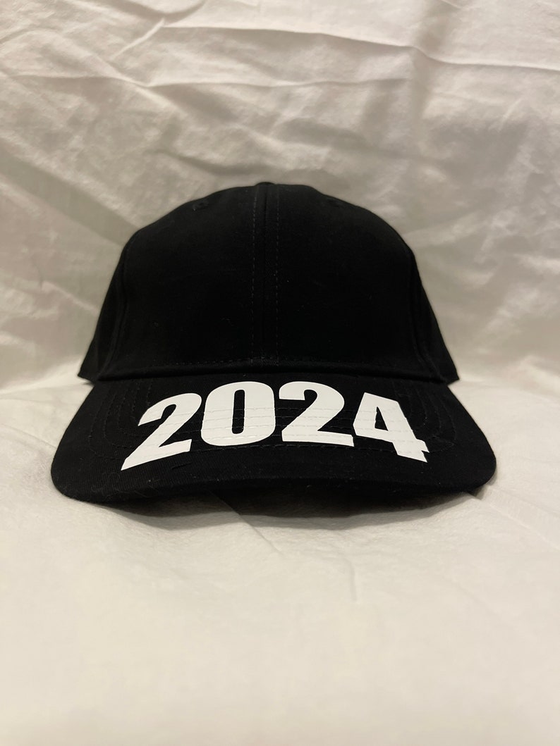 May include: Black baseball cap with the white text "2024" across the front. The cap has a curved brim and a structured crown. The hat is made of a dark fabric.