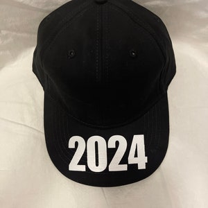 May include: Black baseball cap with the white text "2024" on the front. The cap has a curved brim, a button on top, and stitched details. The hat is made of a solid black material.