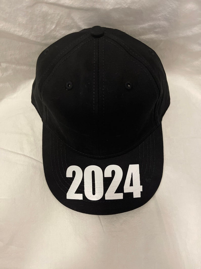 May include: Black baseball cap with the white text "2024" on the front. The cap has a curved brim, a button on top, and stitched details. The hat is made of a solid black material.