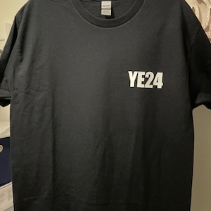 Puede incluir: Camiseta negra de cuello redondo con el texto blanco "YE24" impreso en la parte delantera. La camiseta est&aacute; colgada de una percha blanca. La camiseta est&aacute; hecha de un material suave.