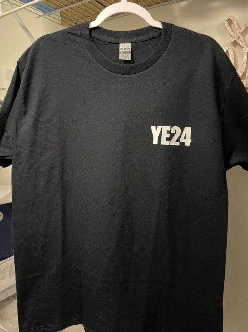 Puede incluir: Camiseta negra de cuello redondo con el texto blanco "YE24" impreso en la parte delantera. La camiseta est&aacute; colgada de una percha blanca. La camiseta est&aacute; hecha de un material suave.