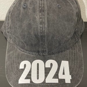 May include: A gray baseball cap with the year 2024 printed in large white block letters on the front. The cap has a curved brim and a classic six-panel design. The fabric appears to be a washed cotton.