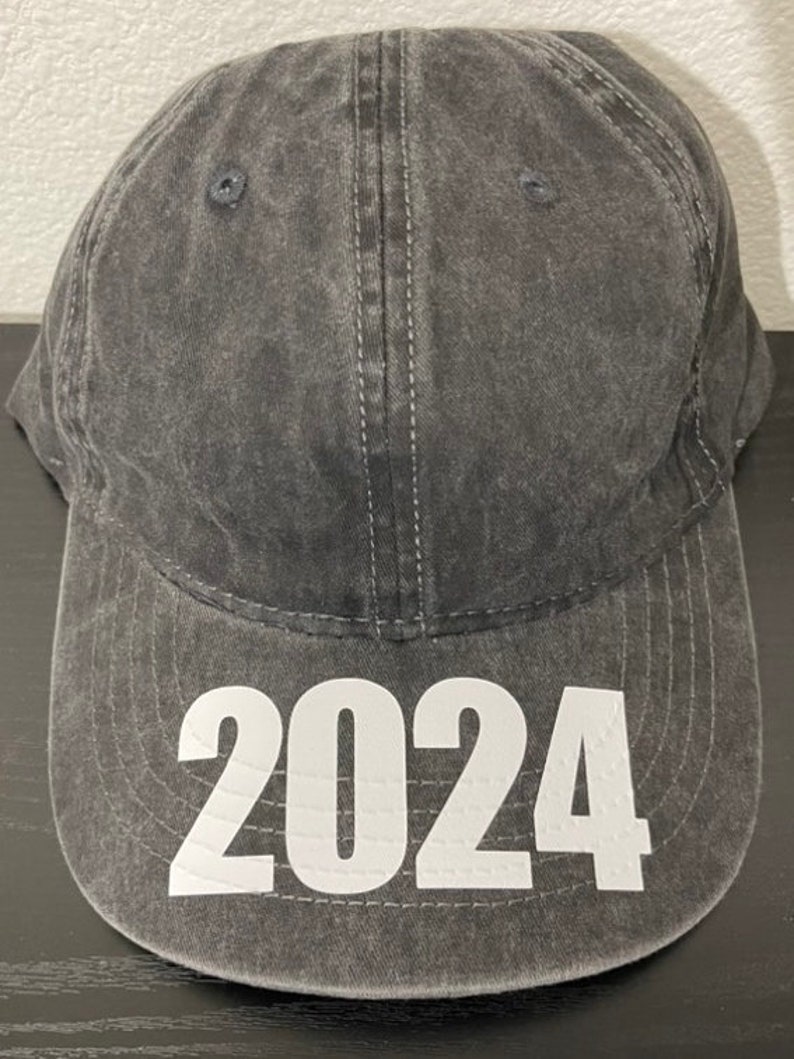 May include: A gray baseball cap with the year 2024 printed in large white block letters on the front. The cap has a curved brim and a classic six-panel design. The fabric appears to be a washed cotton.