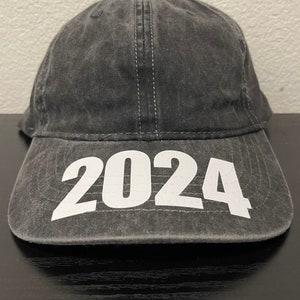 May include: A charcoal gray baseball cap with the year "2024" printed in large white block letters across the front. The cap has a curved brim and a stitched seam down the center.