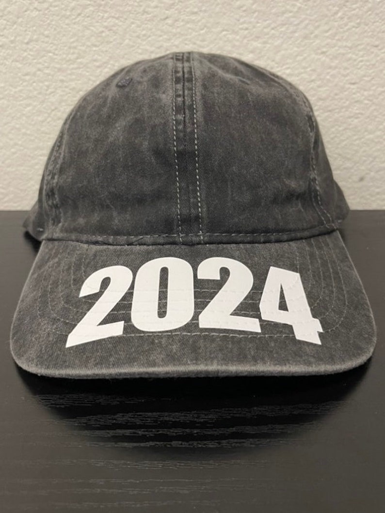 May include: A charcoal gray baseball cap with the year "2024" printed in large white block letters across the front. The cap has a curved brim and a stitched seam down the center.