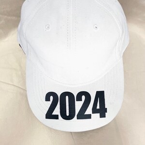 May include: White baseball cap with the black number "2024" printed on the front. The cap has a curved brim, a button on top, and small ventilation holes. The hat is made of a smooth fabric.
