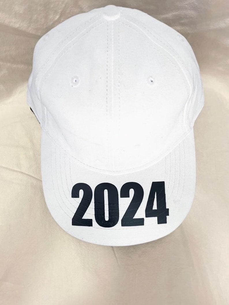 May include: White baseball cap with the black number "2024" printed on the front. The cap has a curved brim, a button on top, and small ventilation holes. The hat is made of a smooth fabric.