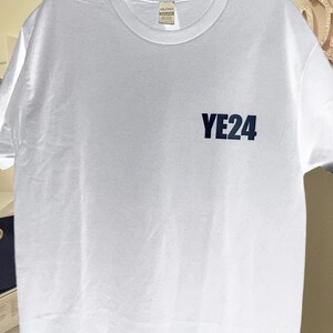Puede incluir: Una camiseta blanca con cuello redondo y mangas cortas. El texto "YE24" est&aacute; impreso en azul oscuro en la parte delantera. La camiseta est&aacute; hecha de un material suave y c&oacute;modo.