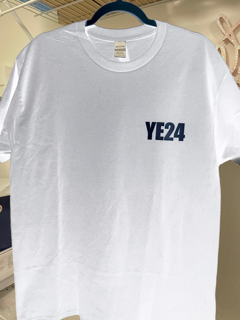 Puede incluir: Una camiseta blanca con cuello redondo y mangas cortas. El texto "YE24" est&aacute; impreso en azul oscuro en la parte delantera. La camiseta est&aacute; hecha de un material suave y c&oacute;modo.