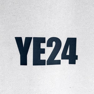 Op de afbeelding: Een close-up van een wit canvas met de zwarte tekst "YE24" gedrukt in een vetgedrukte, schreefloze lettertype. De letters en cijfers zijn gelijkmatig verdeeld en gecentreerd, wat een strakke en moderne esthetiek cre&euml;ert. De textuur van het canvas is zichtbaar.