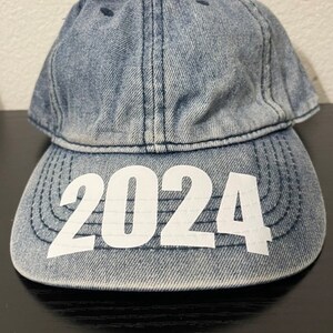 May include: A denim baseball cap with the year "2024" printed in white across the front. The cap is a light blue wash and has a curved brim. The cap is sitting on a black surface.