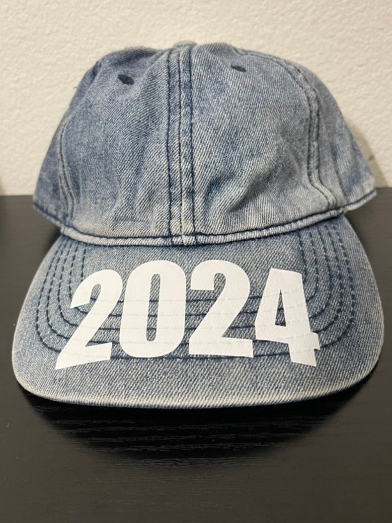 May include: A denim baseball cap with the year "2024" printed in white across the front. The cap is a light blue wash and has a curved brim. The cap is sitting on a black surface.