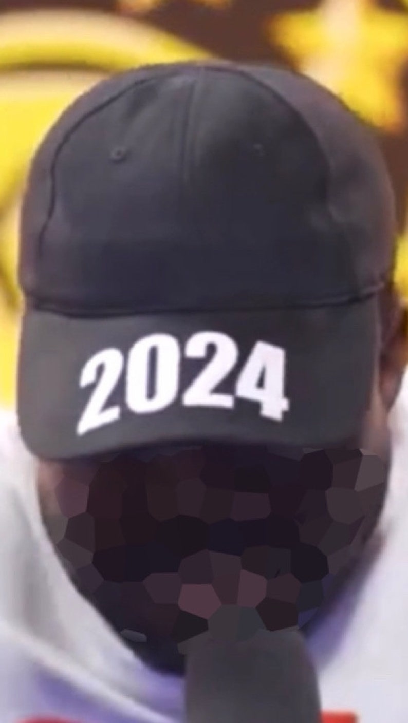 May include: Black baseball cap with the white text "2024" across the front. The cap is a classic six-panel design, with a curved brim and a structured crown. The background is blurred, with yellow and white tones.