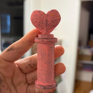 May include: A pink 3D-printed sculpture of a heart on a cylindrical base. The heart and base have visible layered lines, creating a textured look. The sculpture is held in a hand.