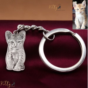 May include: A silver keychain with a small, engraved image of a kitten. The keychain has a silver ring and a twisted silver chain. The image of the kitten is in black and white.