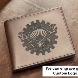 May include: Brown leather wallet with a custom engraved design. The design features a gear, wrenches, and the text "SOB LAKE MECHANICAL EST. 24". The wallet is rectangular and has a stitched border. The text "We can engrave your Custom Logo" is also visible.