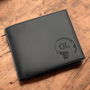 May include: Black leather wallet with a stitched border. The wallet features a fishing-themed design with the initials "GL" and a fish. The wallet is displayed on a wooden surface.
