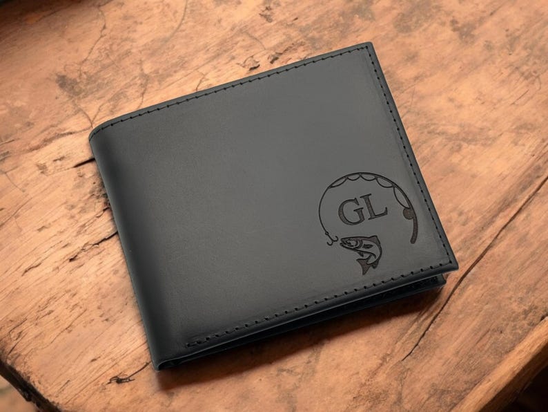May include: Black leather wallet with a stitched border. The wallet features a fishing-themed design with the initials "GL" and a fish. The wallet is displayed on a wooden surface.