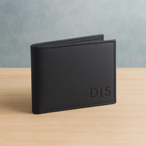 May include: A black leather wallet with the initials "DIS" embossed on the lower right corner. The wallet is rectangular and appears to be bi-fold, with visible stitching around the edges. The wallet is sitting on a light brown wooden surface.