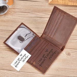 May include: A brown leather bifold wallet featuring multiple card slots and a clear ID window. The interior is engraved with the text 'i love you forever & always Katelyn' accompanied by a heart symbol, matching the text on a white card insert.