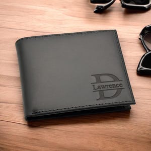 May include: A black leather wallet with the name "Lawrence" and the initial "D" embossed on the front. The wallet is rectangular and has a stitched border. Sunglasses are visible in the background.