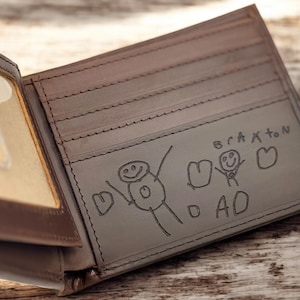 May include: Open brown leather wallet with a clear ID window. The interior features a drawing of stick figures and the word "DAD" with the name "Braxton" above the drawing. The wallet is on a wooden surface.