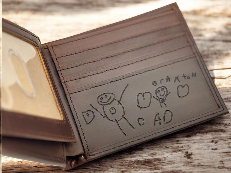 May include: Open brown leather wallet with a clear ID window. The interior features a drawing of stick figures and the word "DAD" with the name "Braxton" above the drawing. The wallet is on a wooden surface.