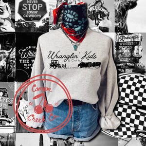Pode incluir: Uma camisola cinzenta clara de gola redonda com o texto preto Wranglin' Kids & Cattle e um gráfico de um veículo utilitário e gado. A camisola é combinada com calções de ganga azuis e uma bandana preta com padrão à volta do pescoço.