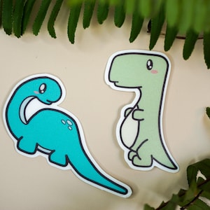 May include: Two cartoon dinosaur stickers, one green and one blue. The green dinosaur is standing upright with its arms crossed. The blue dinosaur is lying down with its head turned to the side. Both dinosaurs have cute, cartoon eyes.