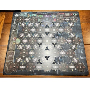 May include: A board game with a dark blue and grey design. The game board features hexagonal spaces labelled with letters A, B, and C, along with question marks. The board has a grid-like pattern with various symbols and text.