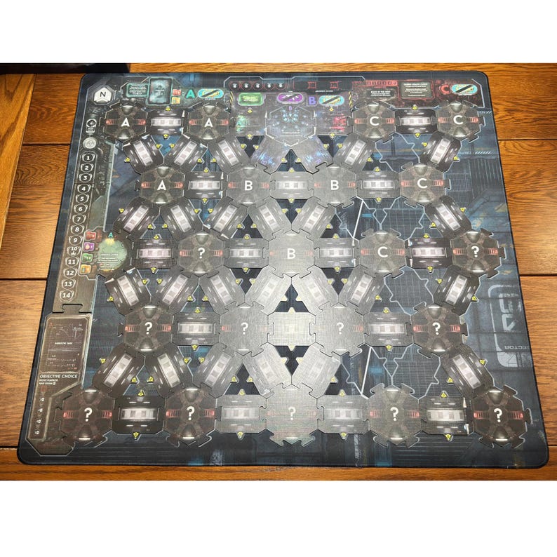 May include: A board game with a dark blue and grey design. The game board features hexagonal spaces labelled with letters A, B, and C, along with question marks. The board has a grid-like pattern with various symbols and text.