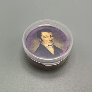 Op de afbeelding: Een ronde, doorzichtige plastic container met een deksel, die een klein, rond, paars en gouden plaatje bevat van een man in een donker pak en witte kraag. De man heeft donker haar en een serieuze uitdrukking.