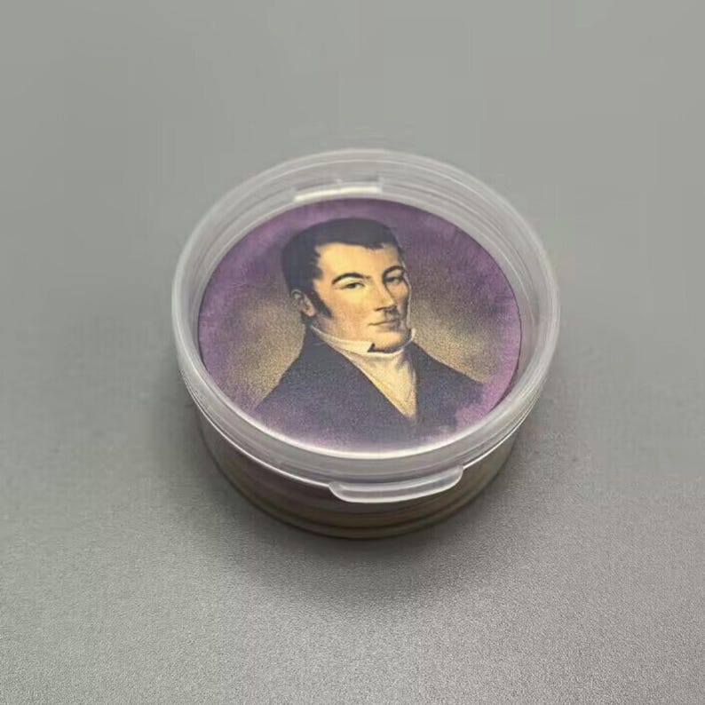 Op de afbeelding: Een ronde, doorzichtige plastic container met een deksel, die een klein, rond, paars en gouden plaatje bevat van een man in een donker pak en witte kraag. De man heeft donker haar en een serieuze uitdrukking.