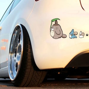 May include: A white car with a decal of Totoro and other characters from the movie My Neighbor Totoro. The decal is on the back of the car, near the rear bumper.