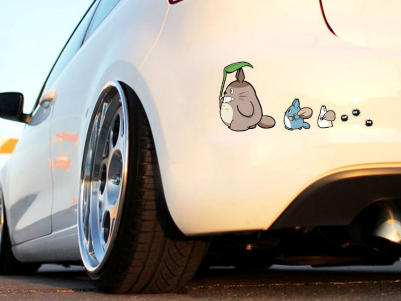 May include: A white car with a decal of Totoro and other characters from the movie My Neighbor Totoro. The decal is on the back of the car, near the rear bumper.