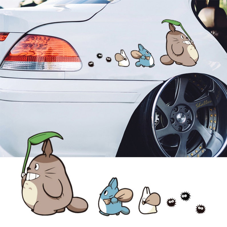 May include: A white car with a decal of cartoon characters from the movie My Neighbor Totoro. The characters include Totoro, a small white bunny, and a blue cat-like creature. The decal is on the back of the car, near the rear wheel.
